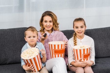 Mother and two children on couch with tubs of popcorn and a remote control.